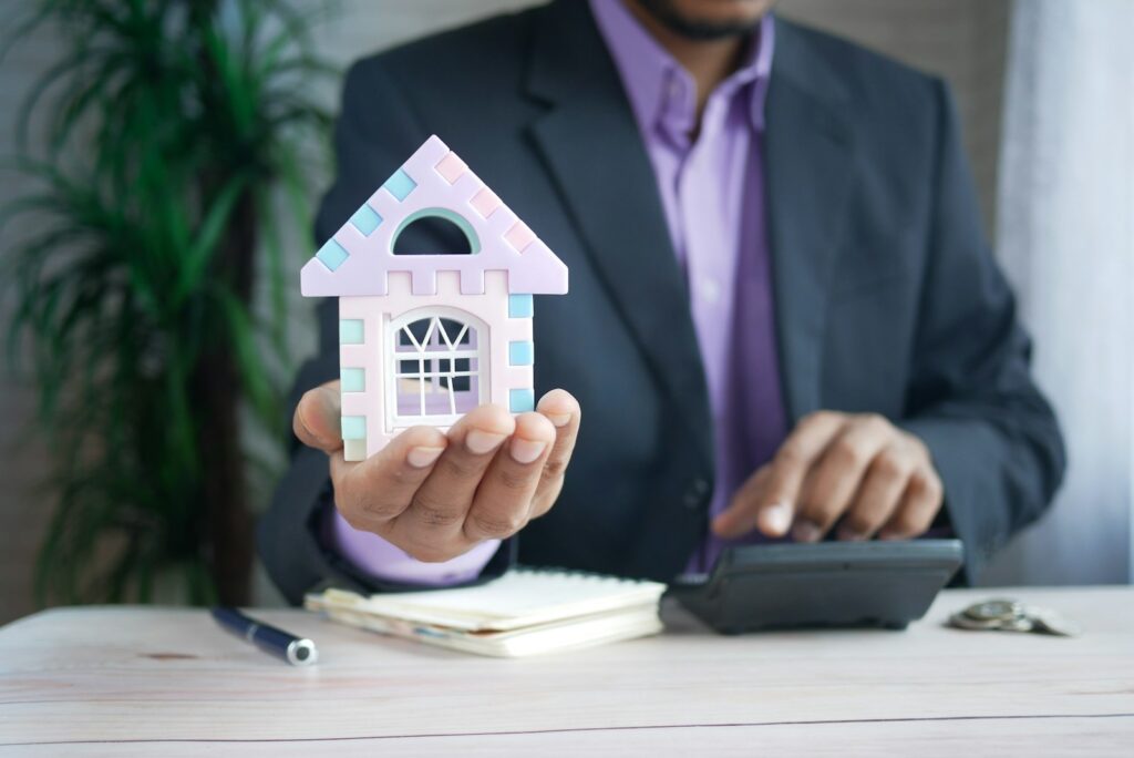 Immobilienmakler hält ein Hausmodell am Schreibtisch neben einem Taschenrechner